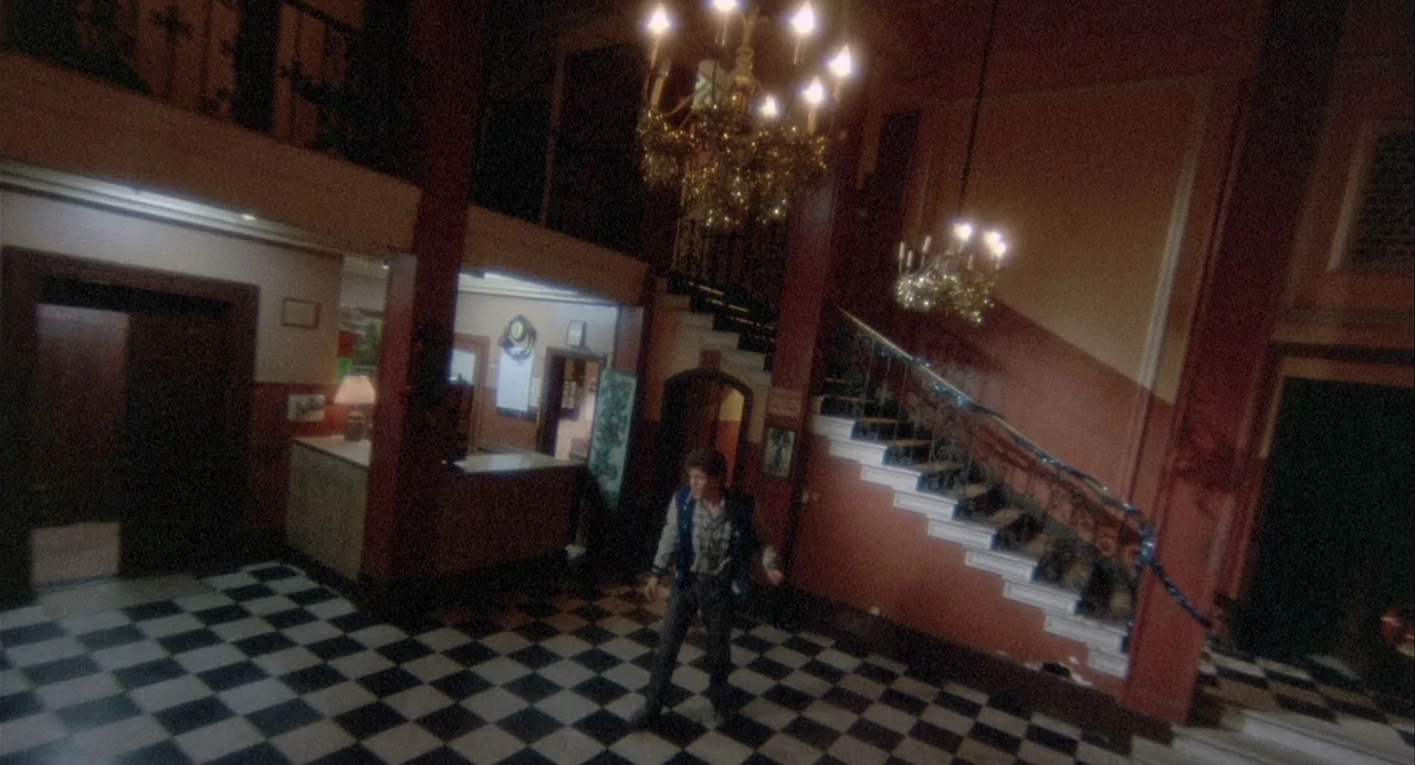 Vamp (1986) movie still: wide — A spacious hotel lobby features dark red walls and a black-and-white checkered floor. A man stands in the center of…