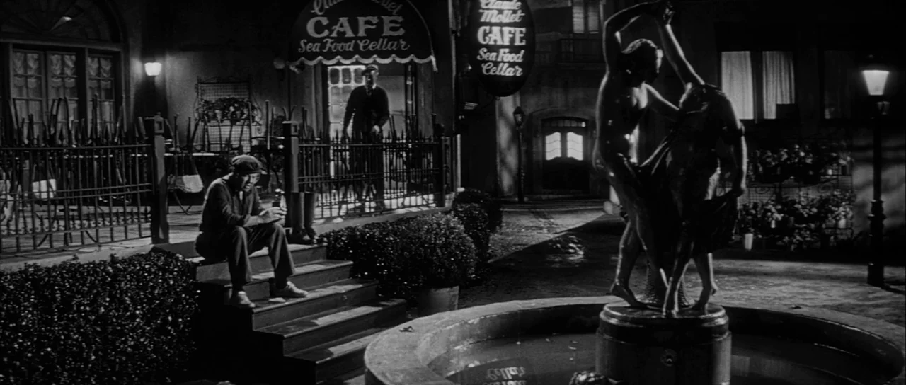 The Tarnished Angels (1957) movie still: wide — A night time city square features a central fountain topped with a stone statue. A man sits on…
