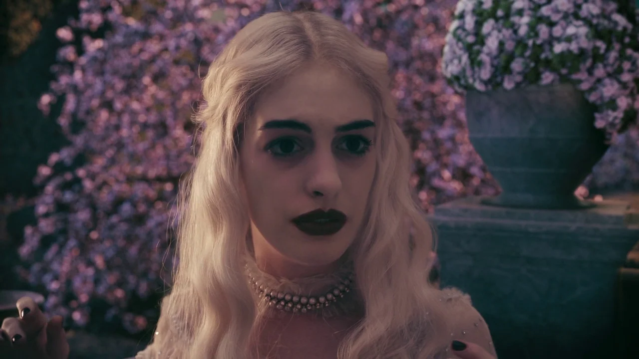 Alice in Wonderland (2010) movie still: closeup — A woman with platinum hair stands in a garden with purple flowering plants. She is wearing a pearl…