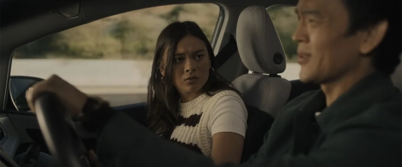 Afraid (2024) movie still: medium — The interior of a moving car shows two people traveling on a highway. A woman in a striped top looks toward the…