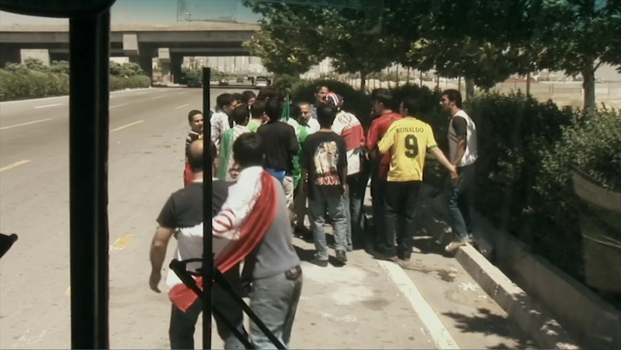 Offside (2015) movie still: medium — A group of men congregates on the dusty shoulder of a wide multi-lane highway. The men face away from the…