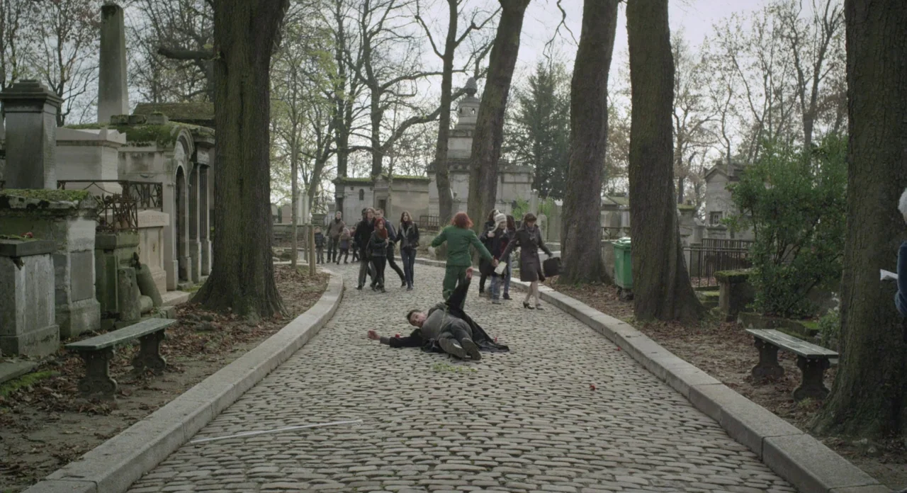 Holy Motors (2012) movie still: wide — A stone-paved walkway winds through a historic cemetery with large trees and stone monuments. A person is…