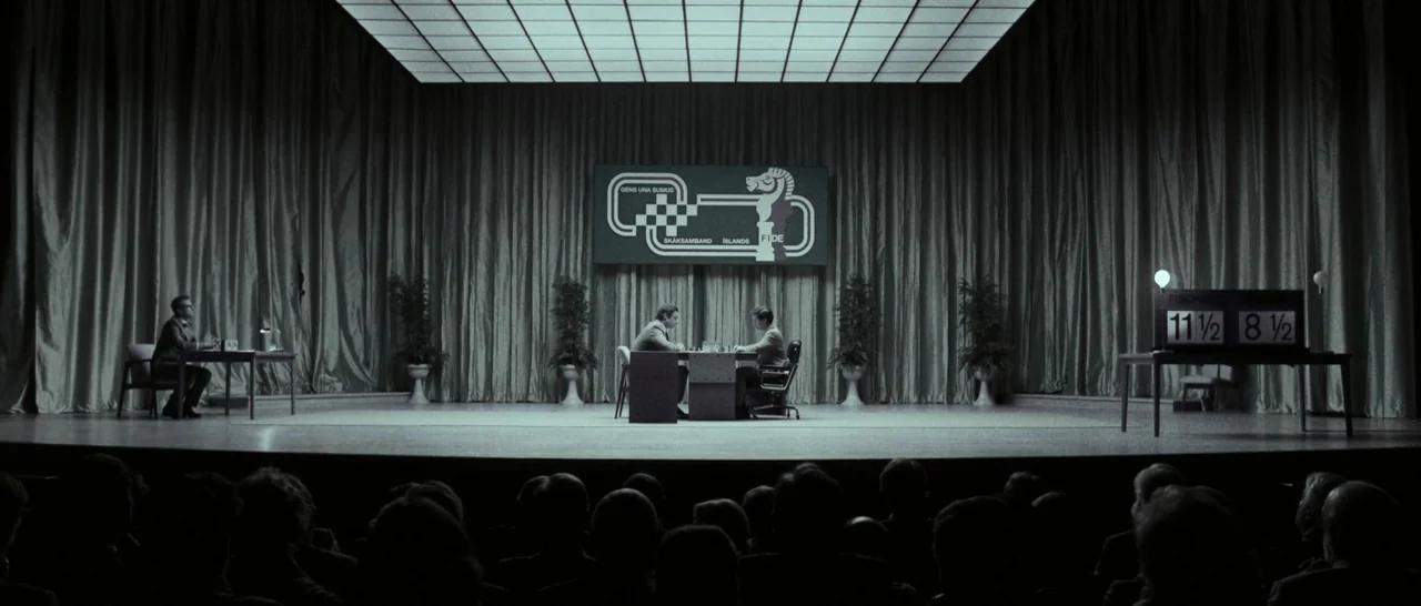 Pawn Sacrifice (2014) movie still: wide — A wide shot shows a chess match taking place on a brightly lit stage. Two men sit at a central table in…
