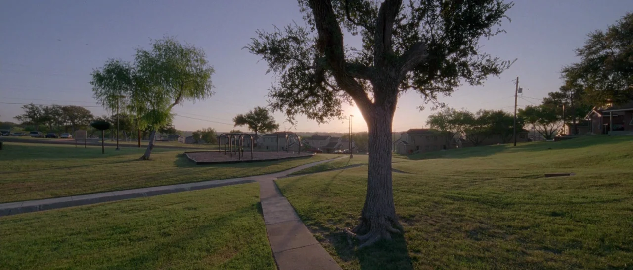 Song to Song (2017) movie still: establishing — A quiet suburban park area features a concrete walkway and a playground structure. A large tree…
