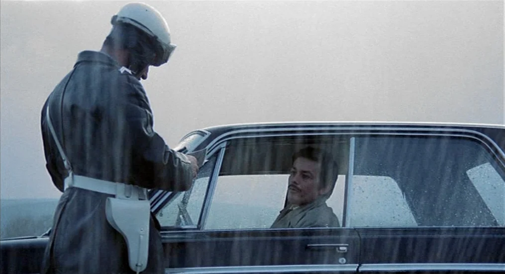 Le Cercle Rouge (1970) movie still: medium — A rain-streaked car window reveals a man sitting in the driver's seat. A police officer in a white…