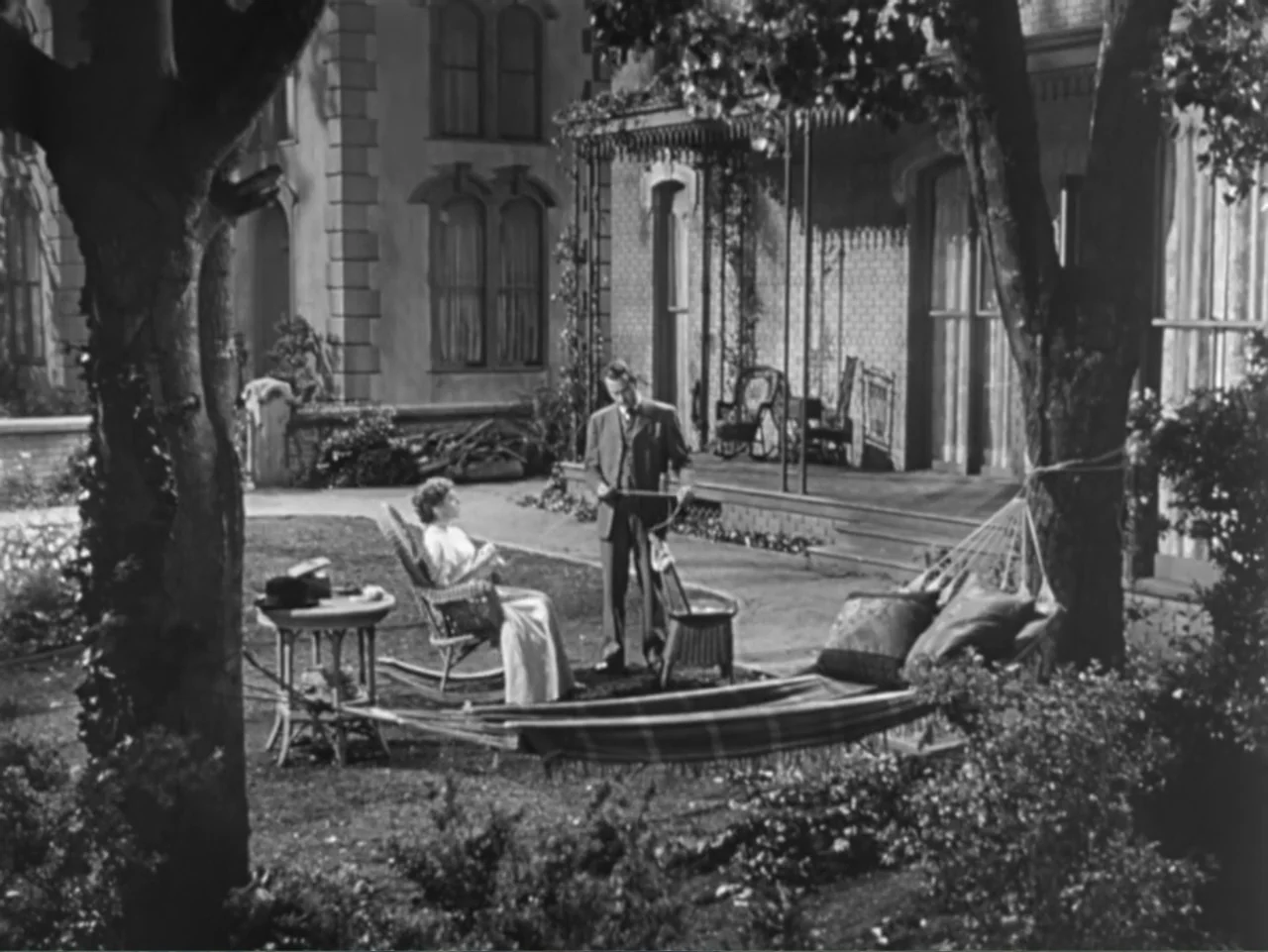 House by the River (1950) movie still: wide — A residential front yard features a porch, a hammock, and mature trees. A man in a suit approaches a…