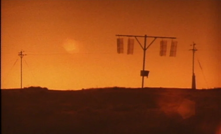 The Garden (1990) movie still: establishing — An orange-hued open field is silhouetted against a setting sun. Three utility structures stand on the…