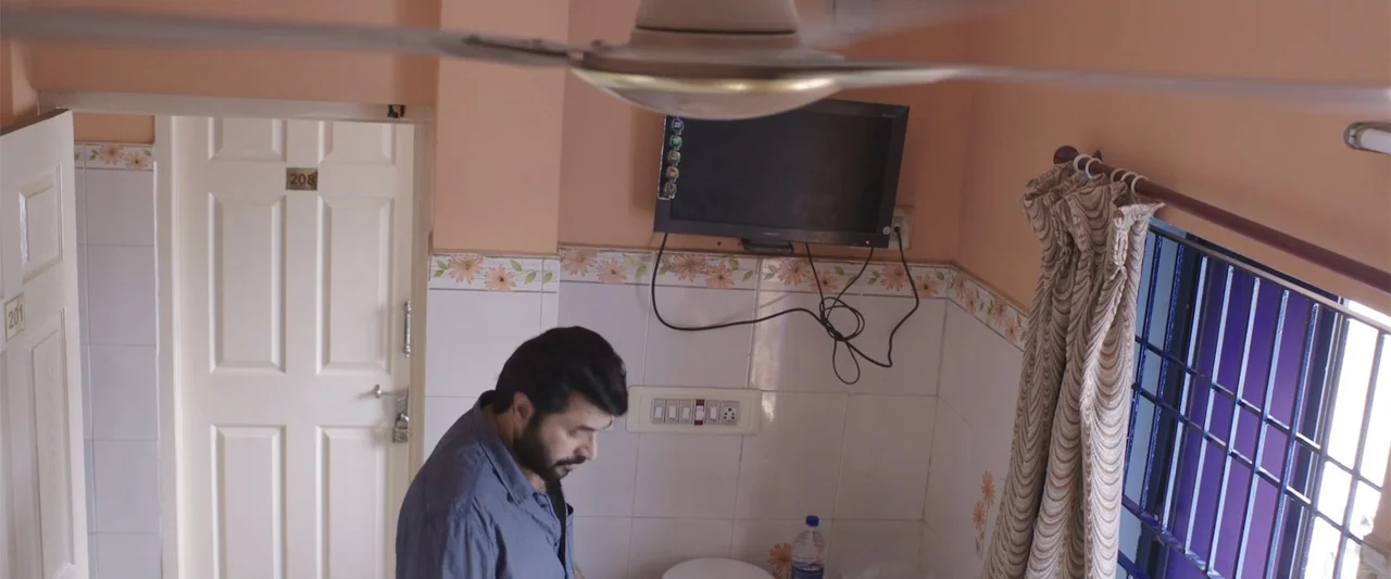 Peranbu (2019) movie still: medium — A small hotel room features white tiled walls and a door labeled 208. A bearded man stands in the foreground…
