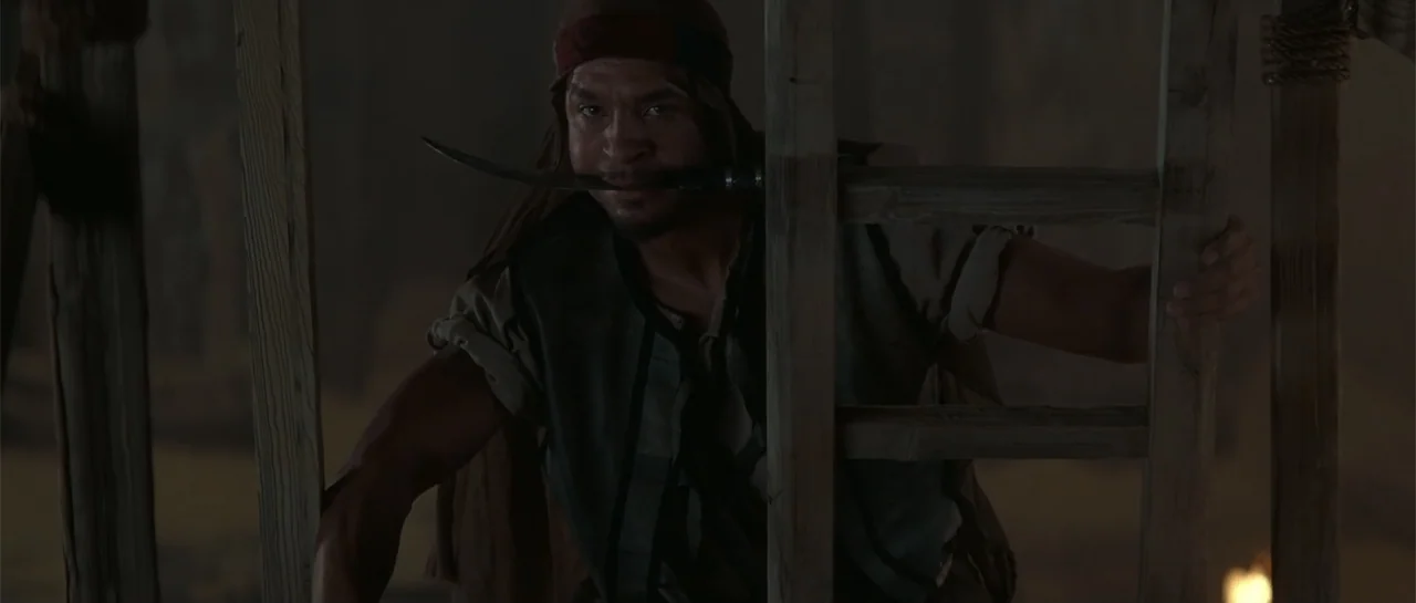 The Mummy Returns (2001) movie still: medium — A man climbs up a wooden structure in a dim, dark environment. He holds a long blade horizontally…