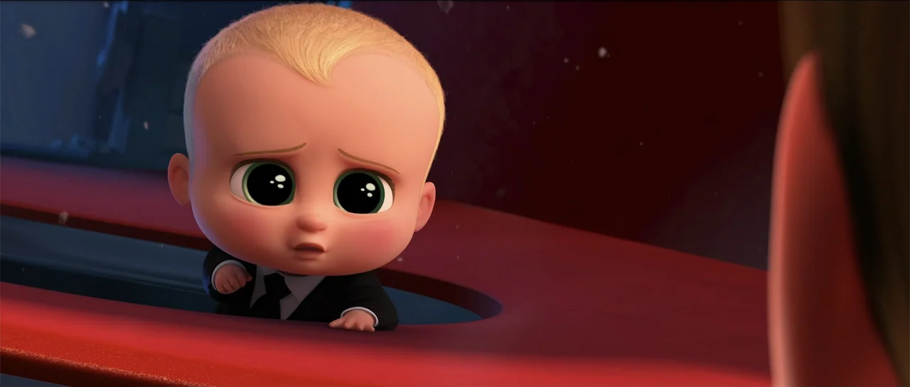 The Boss Baby (2017) movie still: closeup — A close-up of a cartoon baby sitting behind a smooth red desk surface. The baby looks forward with a…