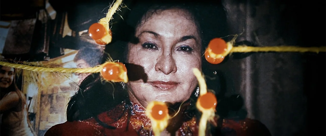 The Kleptocrats (2018) movie still: closeup — A close-up of a printed photograph featuring a woman in a red embroidered shirt. Yellow twine with…
