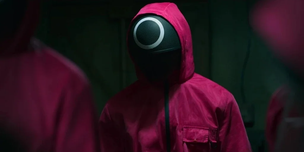Making of: Squid Game (2021) movie still: medium — A dimly lit room with industrial walls contains figures wearing matching magenta jumpsuits. One…