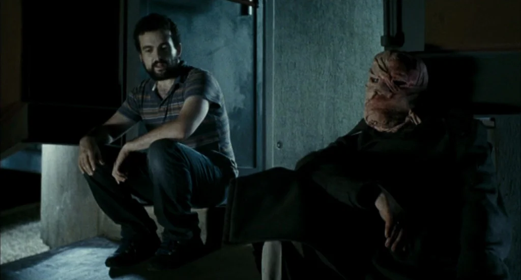 Timecrimes (2007) movie still: medium — A dark stairwell serves as the setting for the two seated figures. A bearded man looks at a person with a…