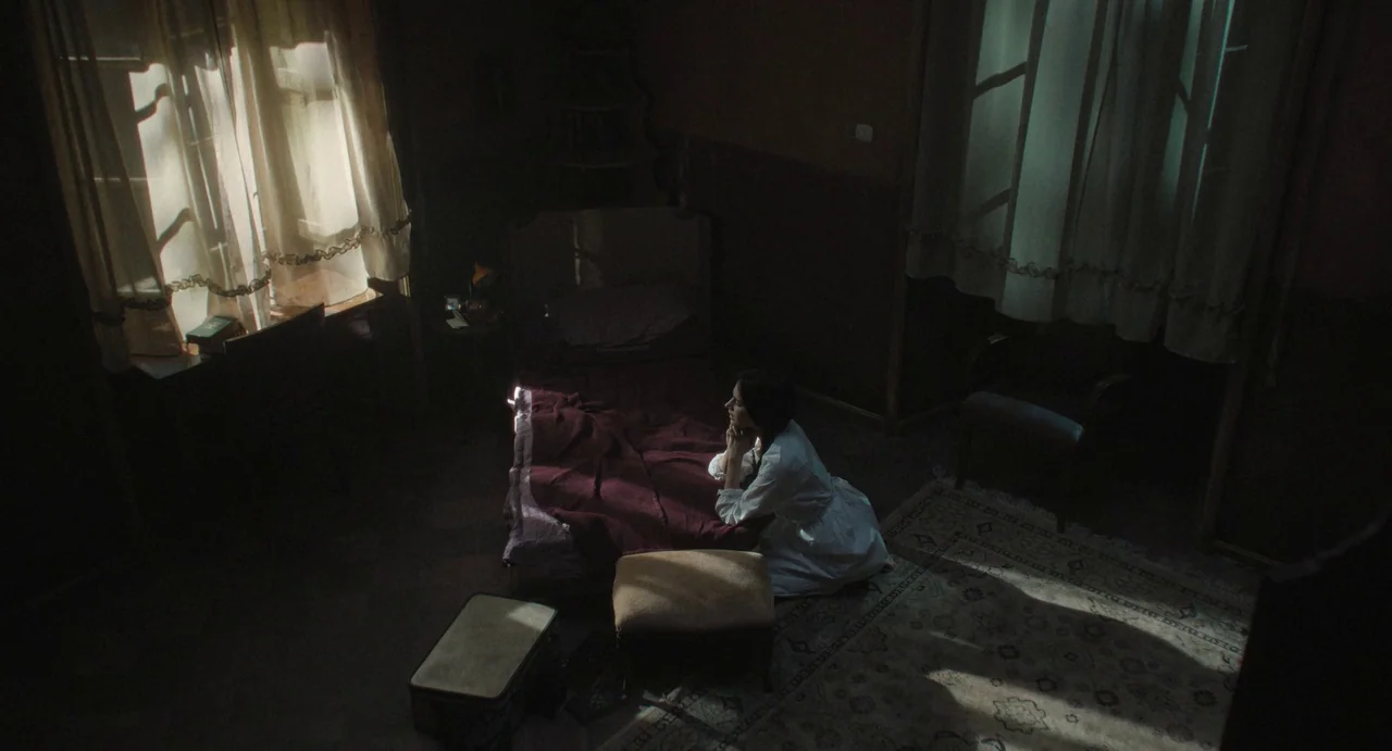The First Omen (2024) movie still: wide — A dimly lit room contains a single bed with a burgundy blanket and a carpet. A woman in a white dress…