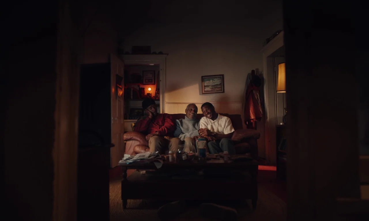 The Last Black Man in San Francisco (2019) movie still: wide — Three men sit on a brown leather couch in a dimly lit domestic space. The room is…
