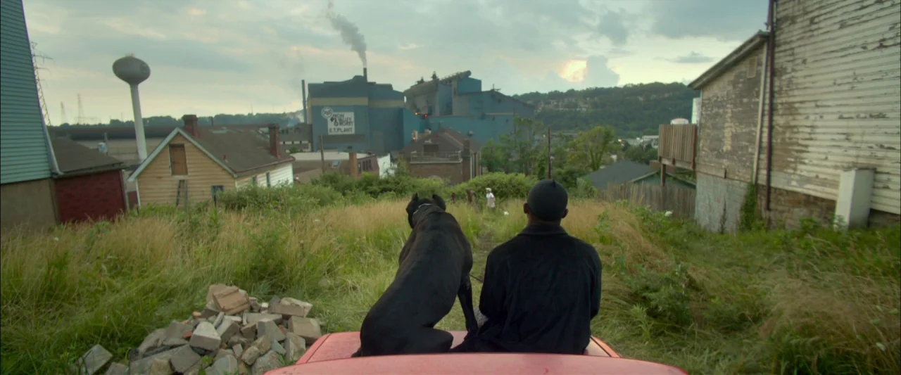 Me and Earl and the Dying Girl (2015) movie still: wide — An overgrown grassy lot faces an industrial plant with tall chimneys emitting white smoke.…