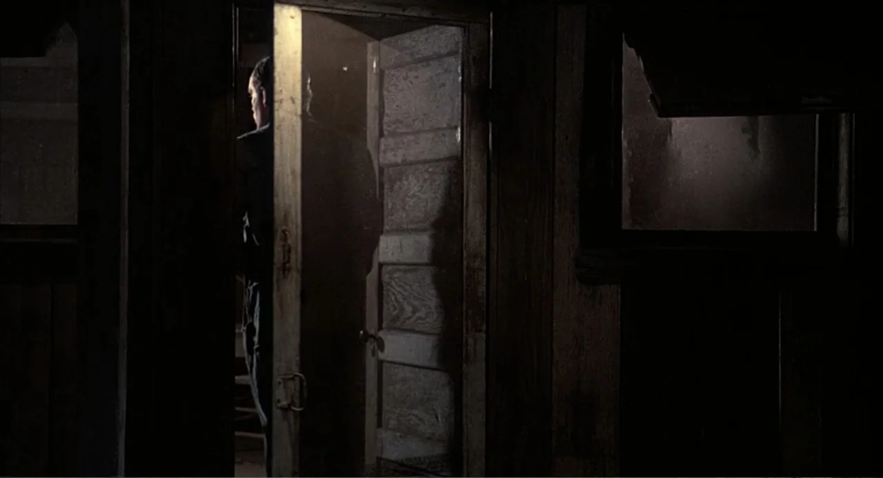 Of Mice and Men (1992) movie still: medium — A dark wooden room interior features an open door revealing a backlit silhouette. A man stands in the…