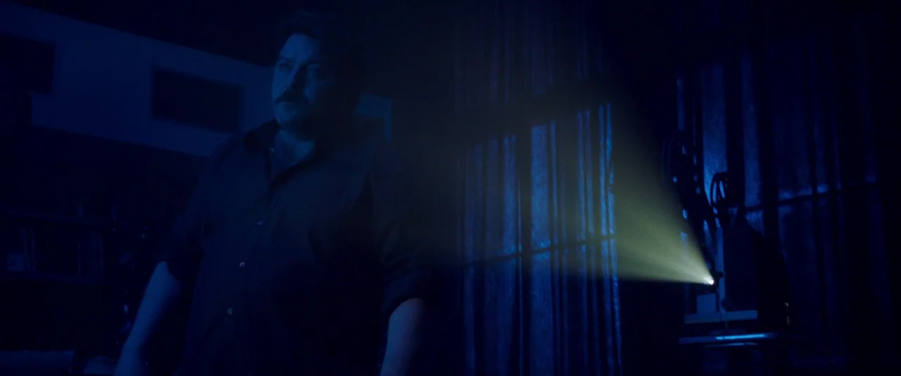 Peter Von Kant (2022) movie still: medium — A dark, minimally furnished room is lit primarily by the glow of a projector. The man stands in the…