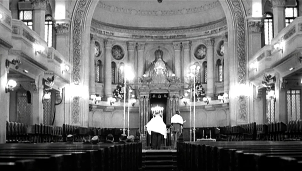 The Tango Lesson (1997) movie still: wide — A spacious synagogue interior features ornate archways and rows of wooden benches. Two men stand at the…