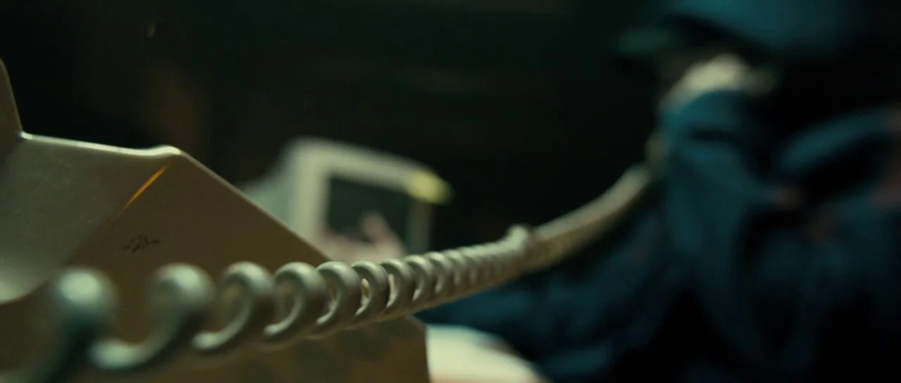 The Imposter (2012) movie still: closeup — A dark space features a vintage telephone receiver and a long coiled cord. The background remains blurry…