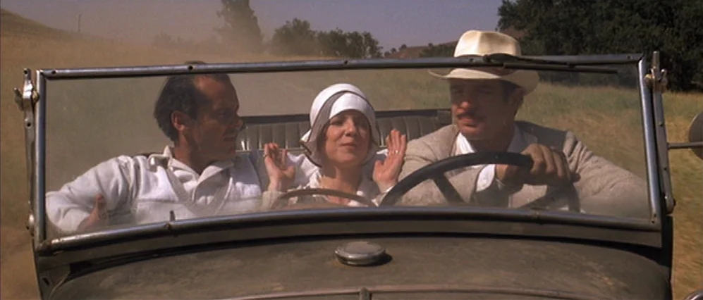 The Fortune (1975) movie still: medium — Three individuals sit side-by-side in a vintage open-top automobile on a dusty rural road. The man on the…