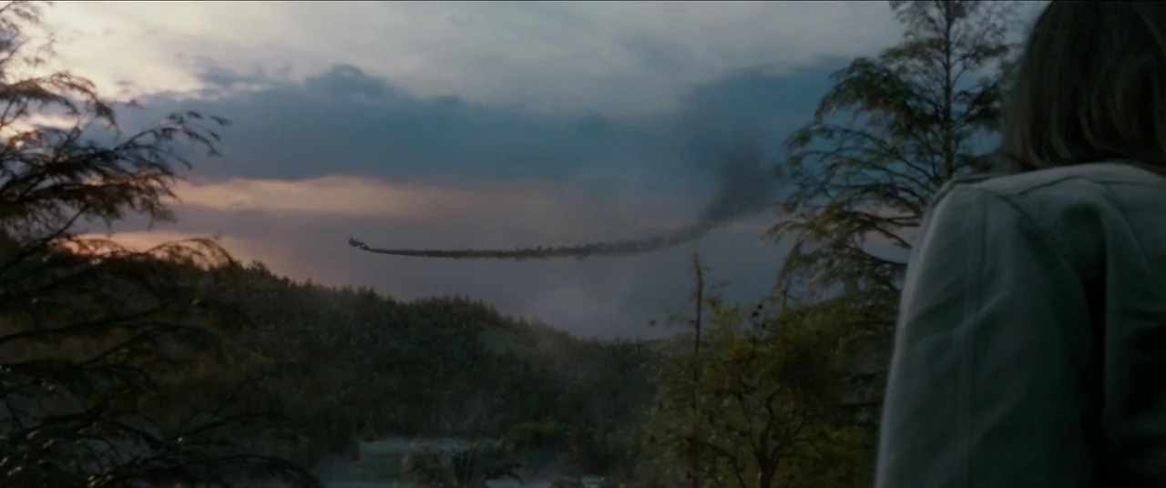 The Predator (2018) movie still: medium — A woman stands in the foreground looking out over a rolling, tree-covered valley. A small aircraft with a…