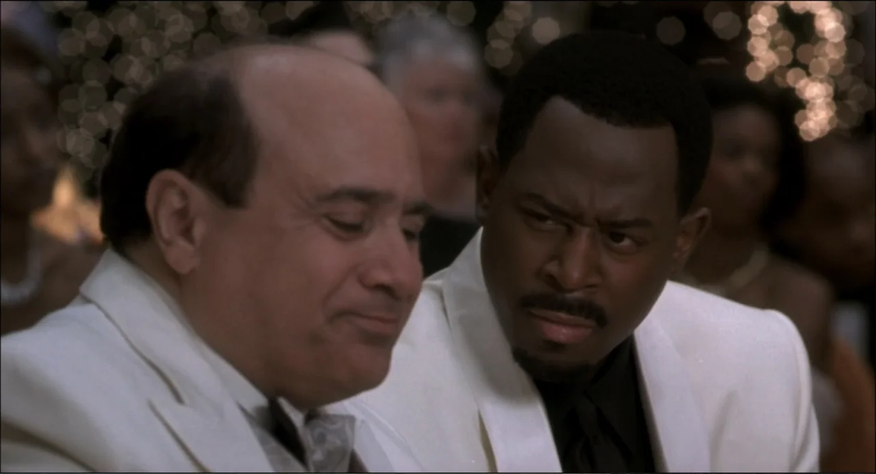 What's the Worst That Could Happen? (2001) movie still: medium — An interior space with a dimly lit crowd visible in the soft-focus background. Two…