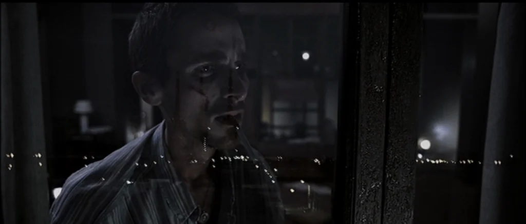 The Machinist (2004) movie still: medium — A dark apartment interior at night viewed through a glass window pane. A thin man with blood on his face…