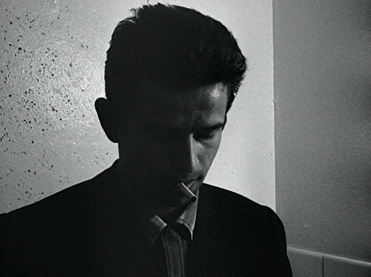 Le Petit Soldat (1963) movie still: closeup — A man stands in a dark corridor with plain wall surfaces. He holds a lit cigarette between his lips…