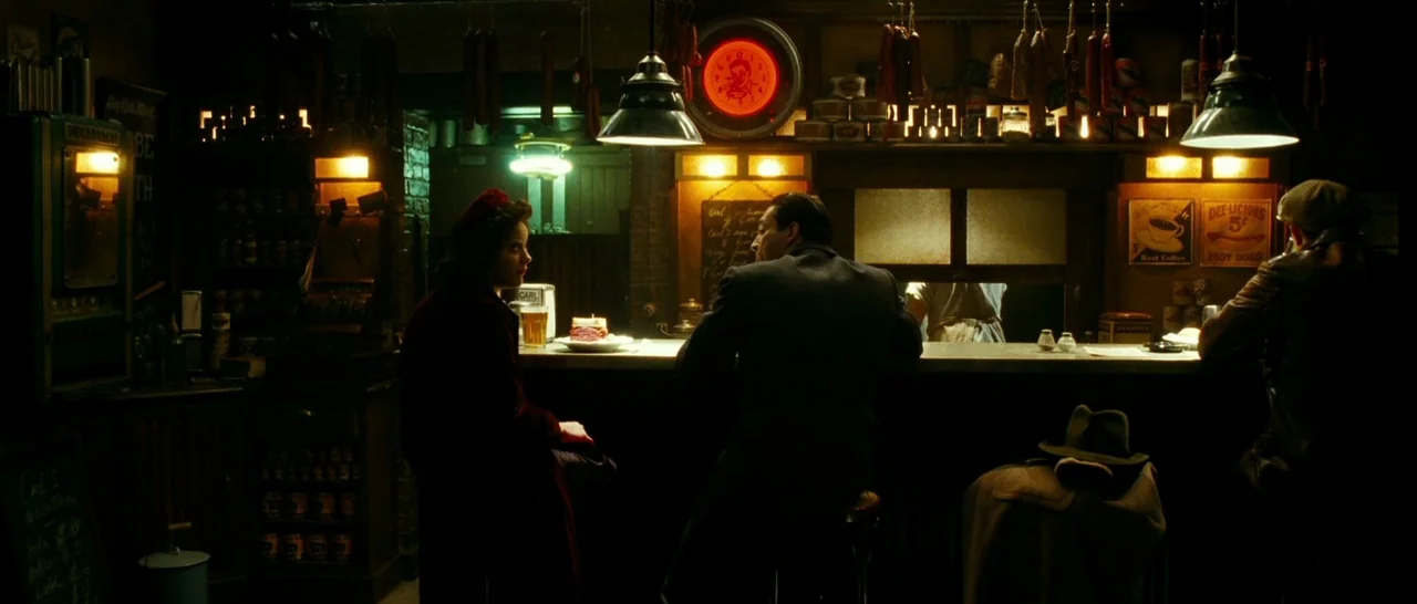 La Vie En Rose (2007) movie still: wide — A dimly lit 1940s-style deli features a long counter with sausages hanging above. Three people sit spaced…