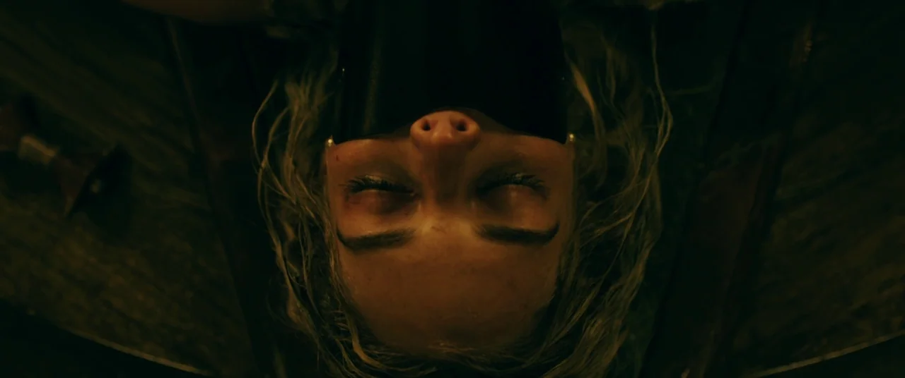 Ready or Not (2019) movie still: closeup — A woman lies flat on a dark, textured wooden surface in a dimly lit space. She has her eyes closed and a…