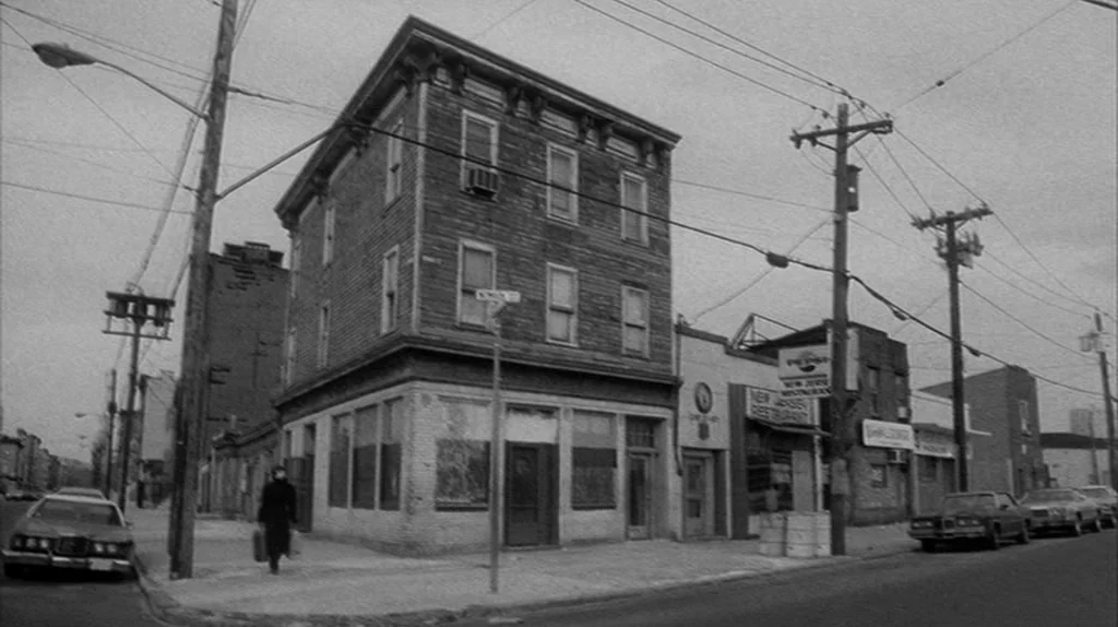Stranger Than Paradise (1984) movie still: wide — A corner building made of brick stands at an urban intersection with power lines overhead. A man…