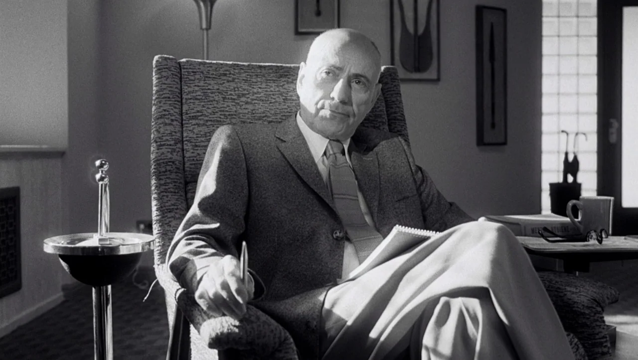 Eros (2004) movie still: medium — A man sits in a padded armchair inside a quiet, sterile professional space. He holds a pen and a notepad while…