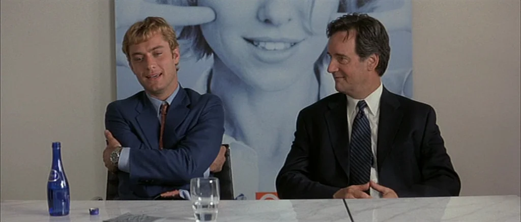 I Heart Huckabees (2004) movie still: medium — A corporate space features a large advertisement showing a face on the wall. Two men in suits sit at…