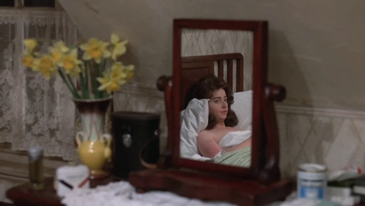 O Lucky Man! (1973) movie still: medium — A bedroom corner features a wooden vanity table with a mirror and yellow flowers. A woman rests in a bed…