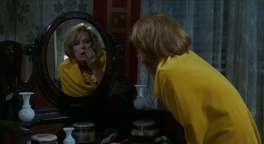 Lisa and The Devil (1973) movie still: medium — A woman stands before a dark wooden dressing table with an oval mirror. She touches her cheek while…