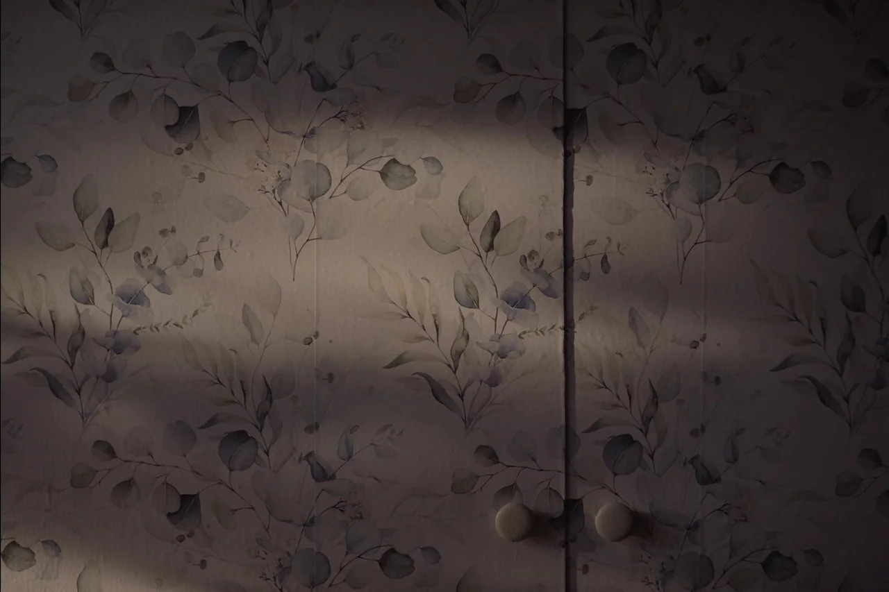 Sometimes I Think About Dying (2023) movie still: closeup — Two cabinet or closet doors are covered in floral wallpaper with muted leaf patterns. A…