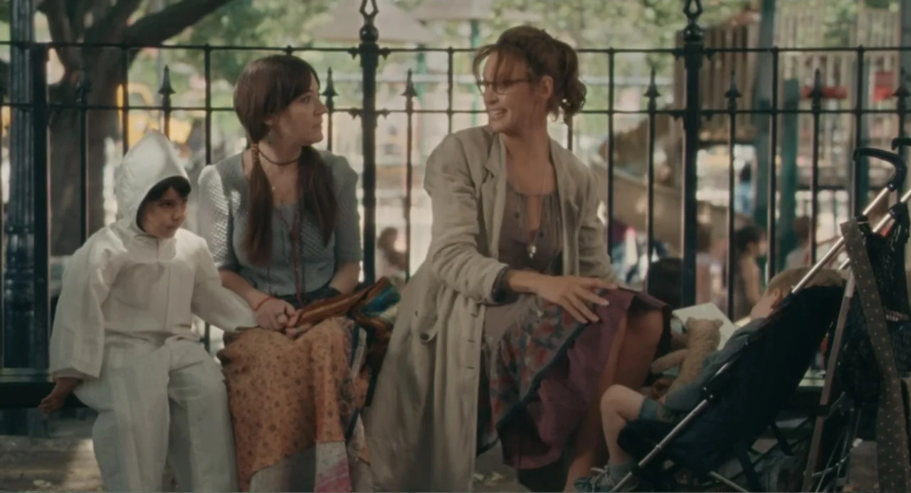 Motherhood (2020) movie still: medium — Two women and a child sit on a bench inside a park. The adults engage in conversation while the child sits…