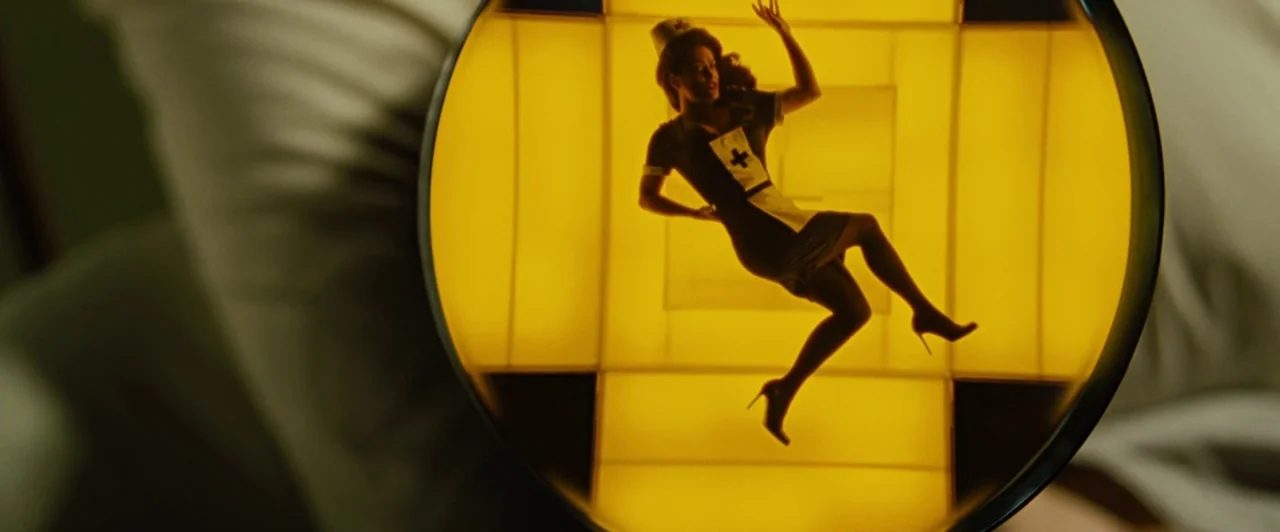 Across the Universe (2007) movie still: closeup — A circular view through a magnifying glass reveals a woman silhouetted against yellow panels. The…