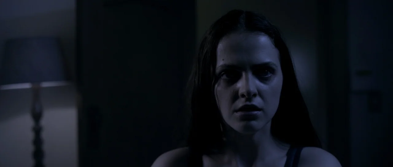 The Lullaby (2019) movie still: closeup — A darkened interior space features a woman standing in front of a lamp. She faces the camera directly with…