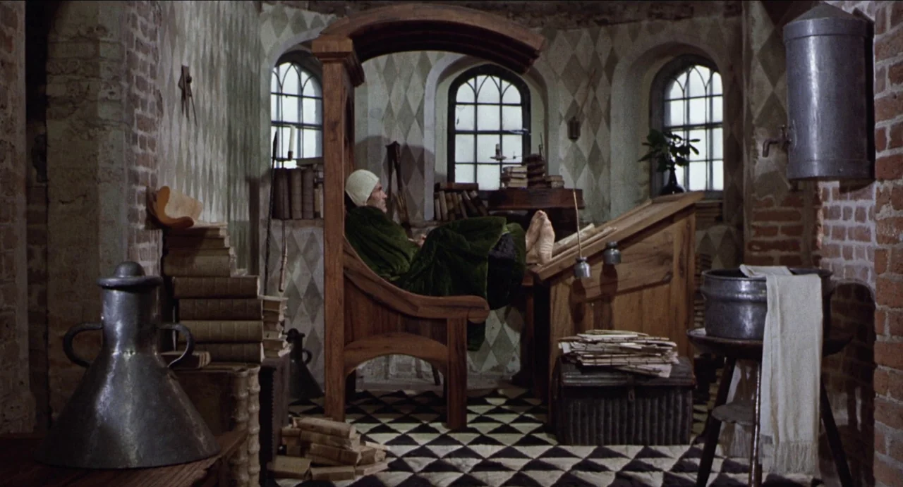 The Canterbury Tales (1972) movie still: medium — A stone study with diamond-patterned walls contains wooden furniture, stacks of books, and…