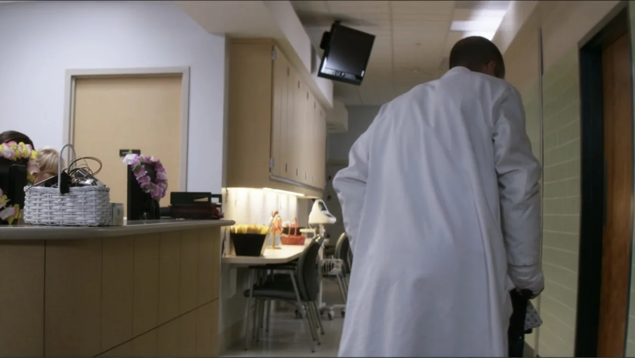 Parker (2013) movie still: medium — A hospital corridor features light green tiled walls and a beige reception desk. A man in a white lab coat walks…