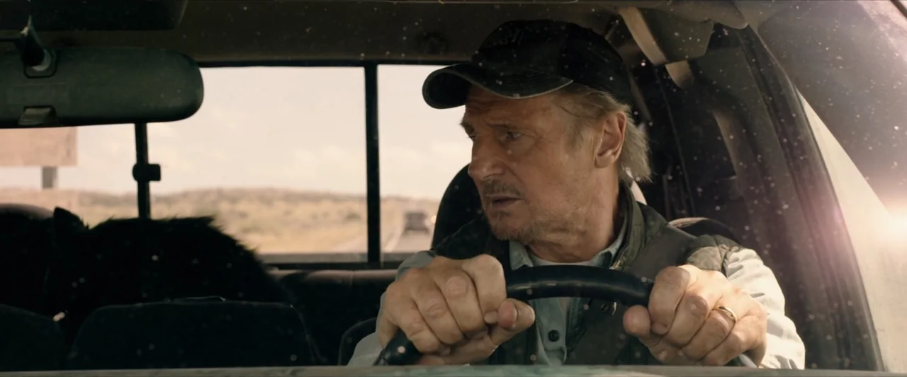 The Marksman (2021) movie still: medium — The interior of a vehicle shows a man driving along a dusty road. He grips the steering wheel while…
