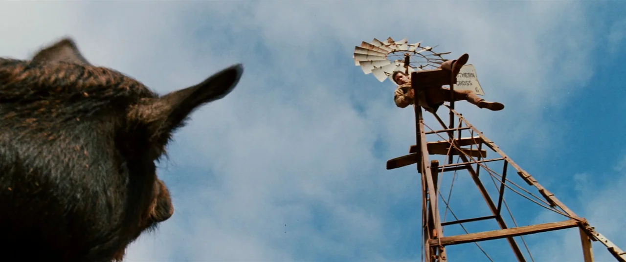 Razorback (1984) movie still: wide — A tall metal windpump stands against a clear, bright blue sky. A young man sits at the top of the tower looking…