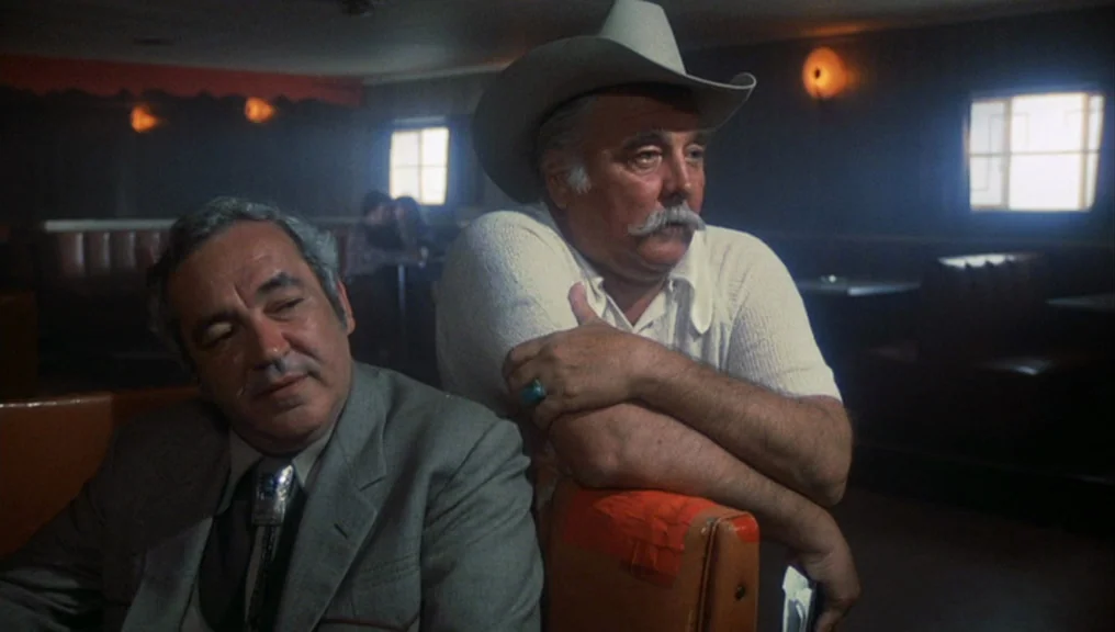 Alice Doesn’t Live Here Anymore (1974) movie still: medium — A dimly lit 1970s diner with dark leather booths and backlit windows. Two men sit…