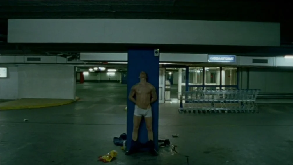 Import Export (2007) movie still: fullbody — A dimly lit parking garage features a central blue structural pillar and discarded litter. A man in…