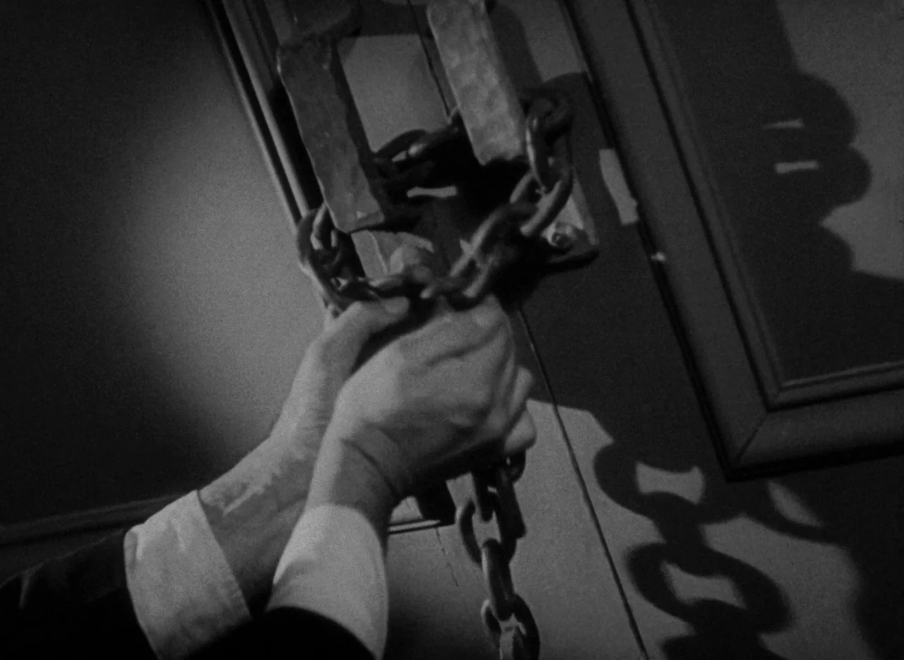 The Invisible Man (1933) (1933) movie still: closeup — A dark interior view of a hand locking a metal chain across a door. The chain is pulled taut…