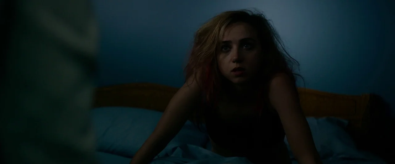 The Monster (2016) movie still: medium — A dimly lit room with a blue-toned wall and a wooden headboard. A young woman with messy hair leans forward…
