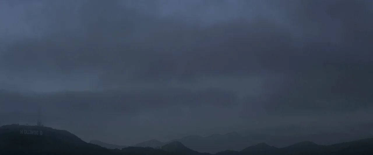 Starry Eyes (2014) movie still: establishing — The Hollywood sign sits on a dark, rolling hillside under a vast, overcast sky. Thick clouds blanket…