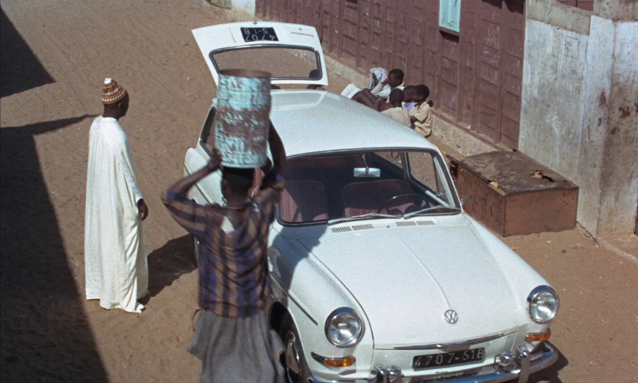 Mandabi (1968) movie still: fullbody — A dusty road in a residential area with a white sedan parked. A person walks past the car carrying a…